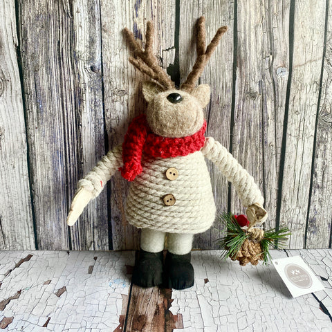 Textile reindeer figure with a red scarf, white sweater, and black boots against a wooden background.