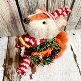 Stuffed fox toy wearing a knitted hat and scarf with a small wreath, on a wooden surface.