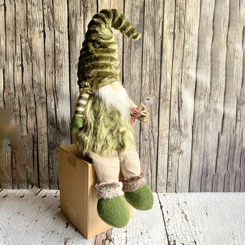 Gnome figurine with green hair and shoes sitting on a cardboard box against a wooden wall.