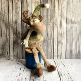 Stuffed moose toy with a green hat and scarf on a wooden block against a wooden wall.